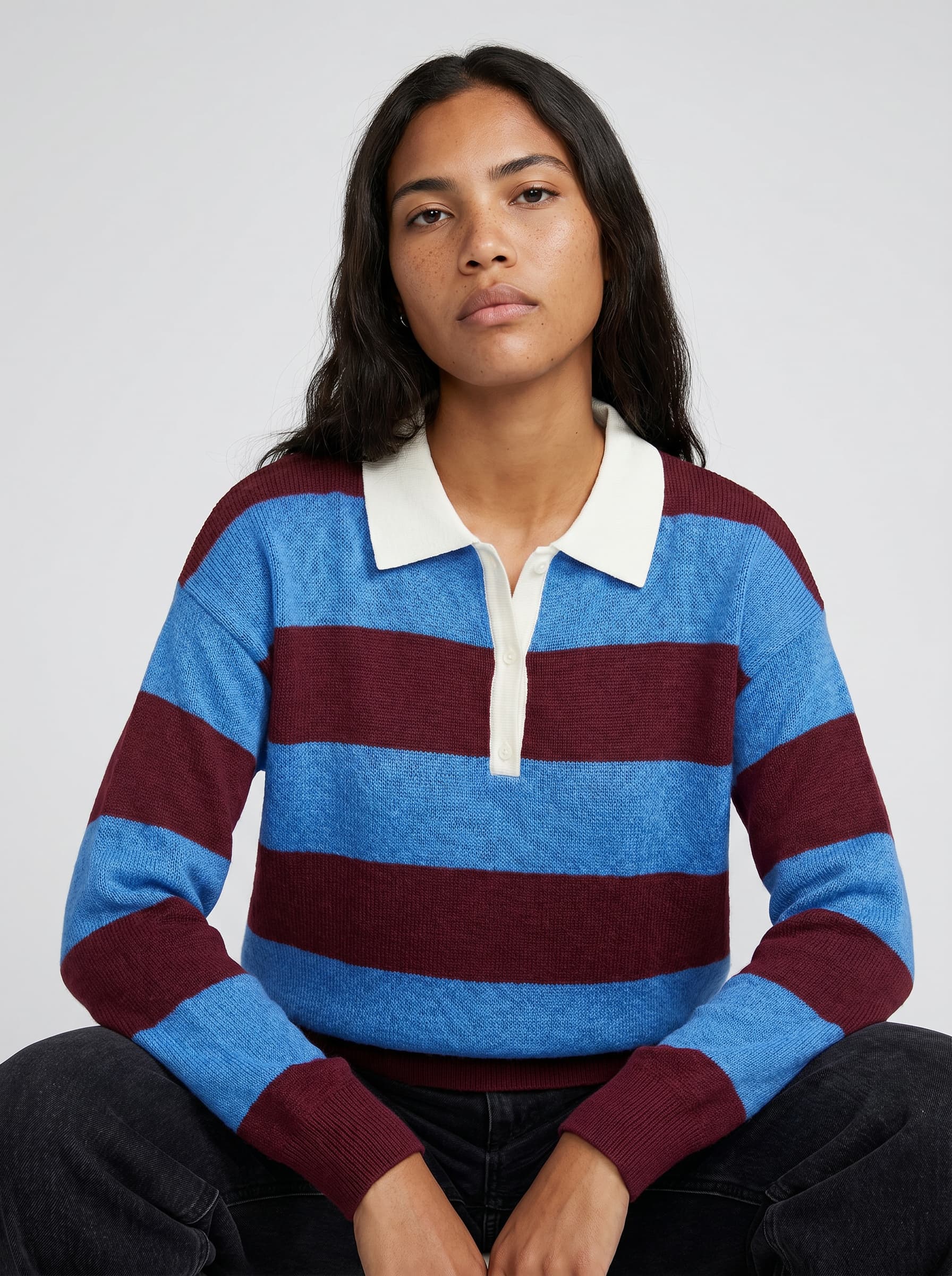 Model in a blue and burgundy striped rugby polo with white collar, seated in black jeans