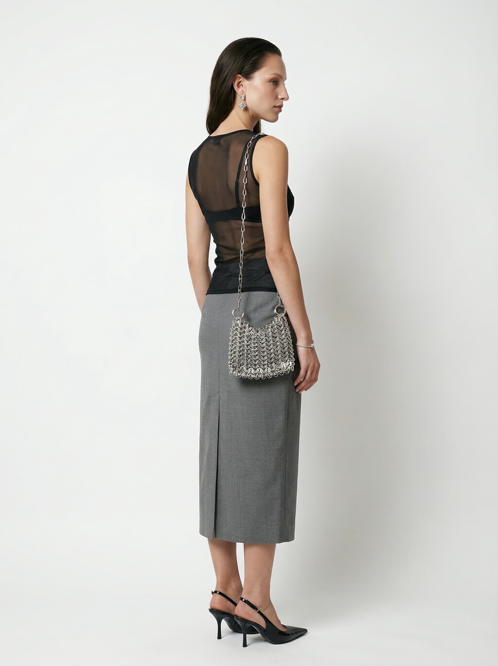 A woman with long, dark hair is wearing a sleeveless, sheer black top and a grey, high-waisted skirt, paired with black heels and a chain-strap silver bag, standing against a plain white background.