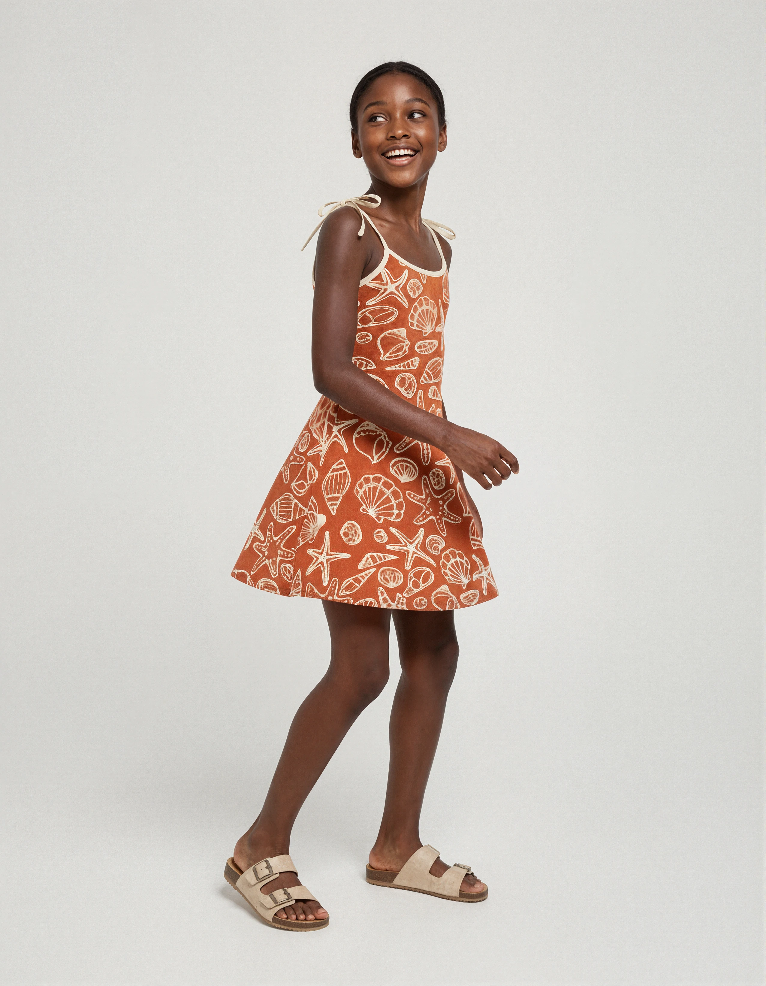 Smiling girl in an orange seashell-print sundress and beige sandals against a light backdrop