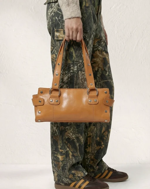 AI model in camouflage cargo trousers holding a tan studded leather shoulder bag, brown sneakers on a white studio floor