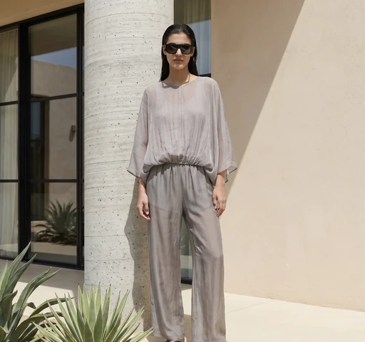 Model in a sheer grey wide-sleeve blouse and trousers with black sunglasses standing by a concrete pillar outdoors