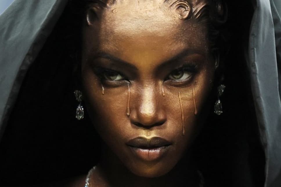 Close-up of a hooded model with gold-tinted skin, drop earrings and painted tears on the Met Gala red carpet