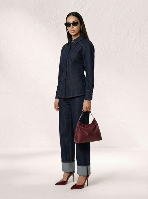 A person stands against a textured white background, wearing a stylish dark denim jacket and matching jeans, accessorized with black sunglasses, burgundy high heels, and holding a maroon handbag, combining classic denim fashion with chic accessories.