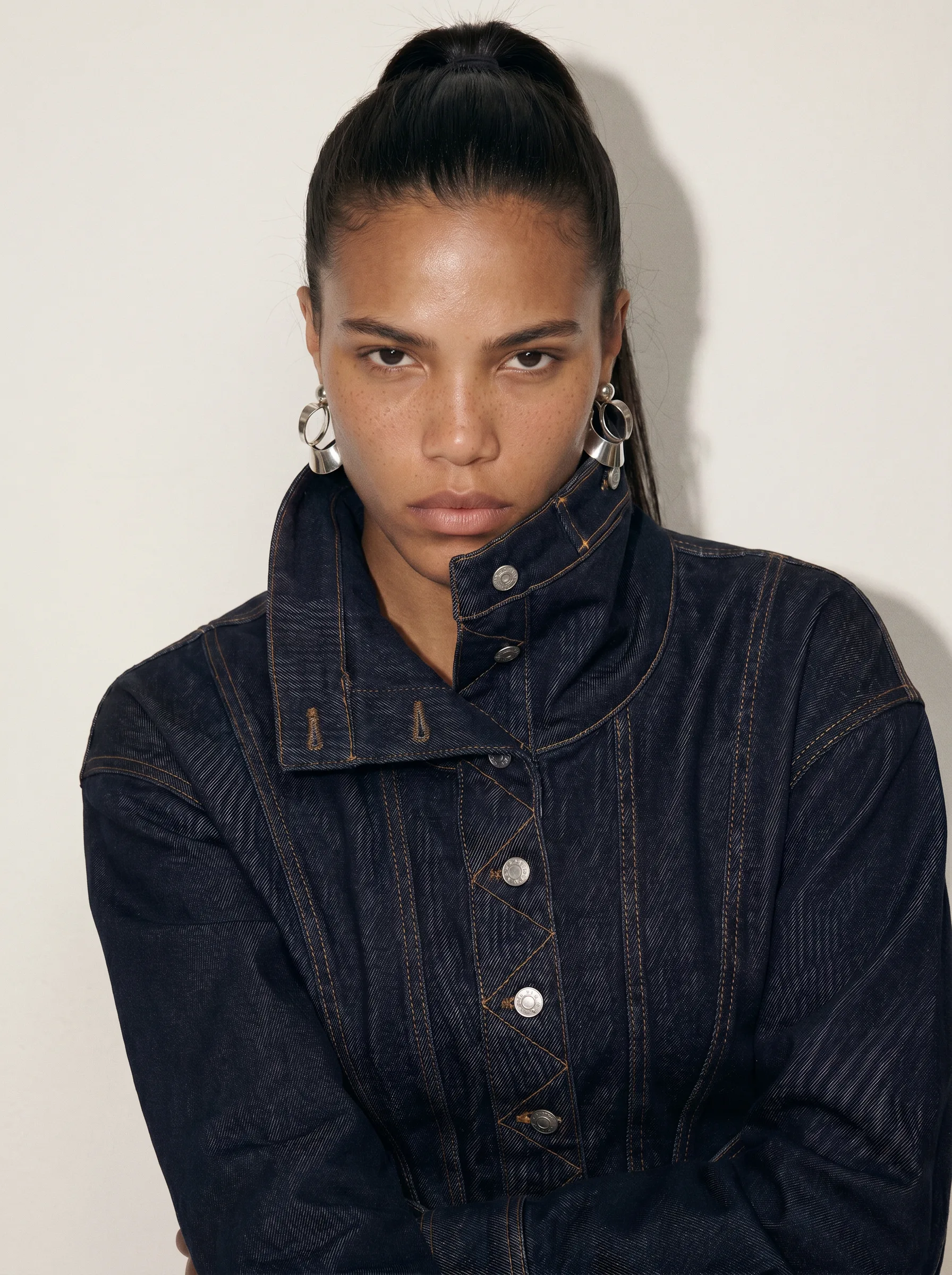 Model with a slicked-back ponytail and silver hoop earrings in a high-collared dark denim jacket, arms crossed