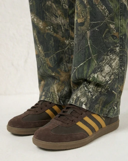 Close-up of camouflage-print cargo trousers over brown suede sneakers with gold stripes on a white studio floor