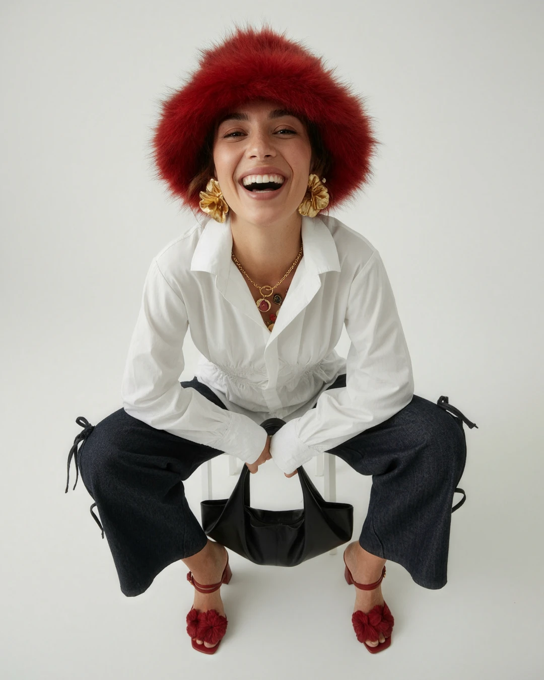 Laughing model in a red faux-fur hat, white shirt and dark tie-side jeans crouching with a black bag and red sandals