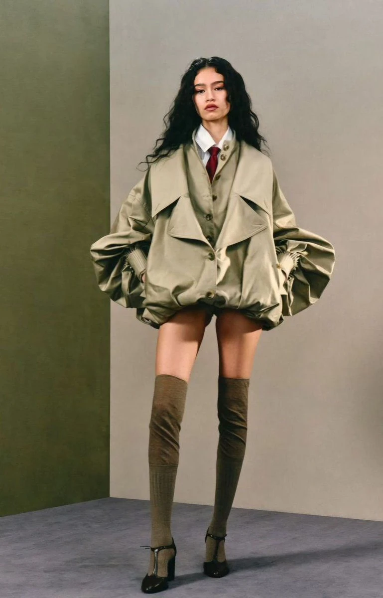 Model with long curly hair in a khaki balloon-hem trench, red tie and over-the-knee socks against a two-tone wall