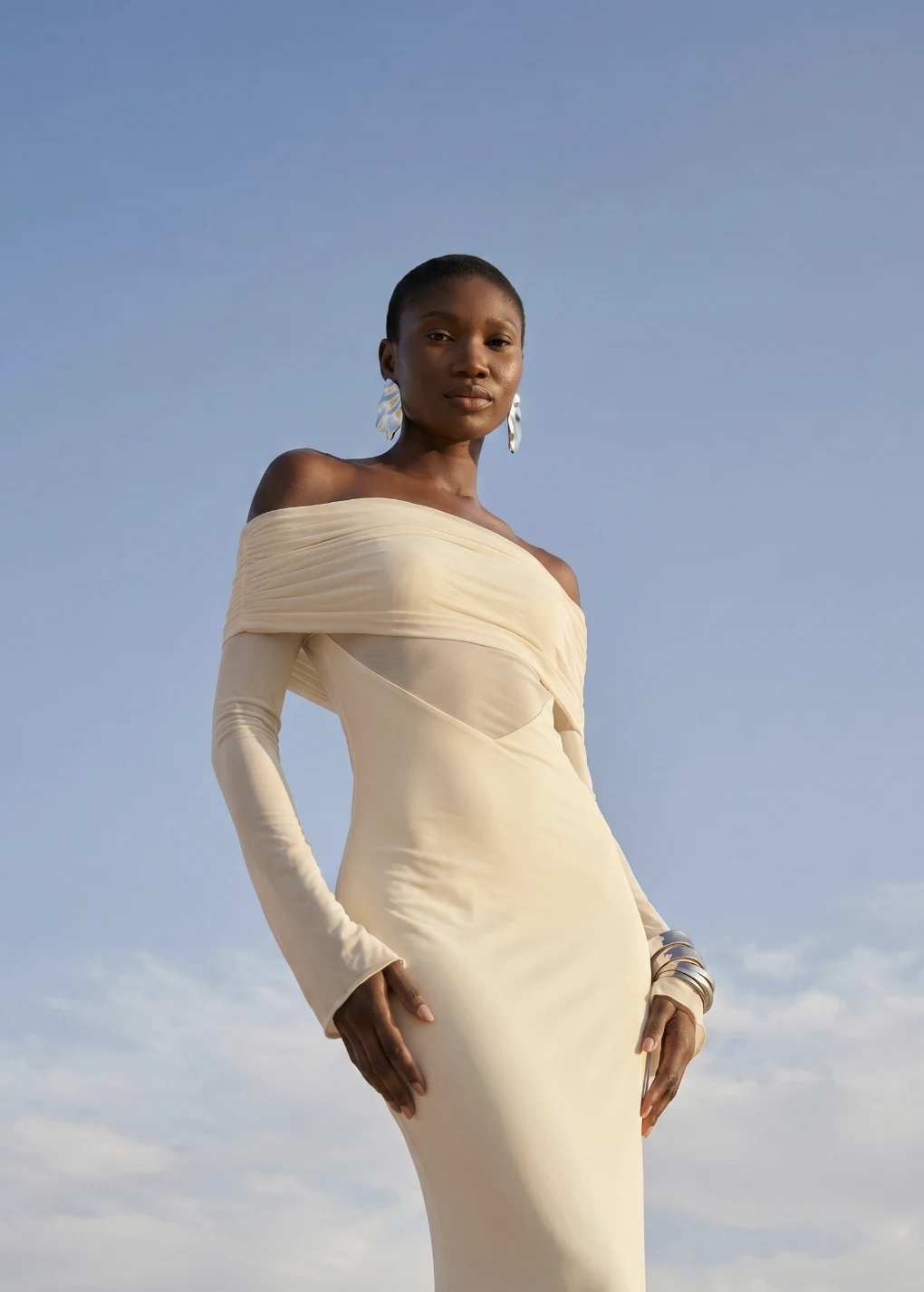 AI model in a cream off-shoulder dress with silver earrings against a blue sky, generated with Veeton