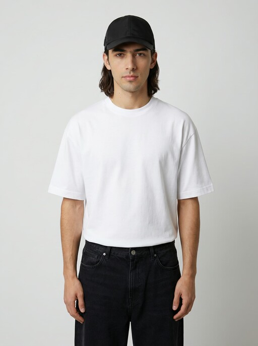 A person wearing a plain white t-shirt and a black cap stands against a neutral gray background, looking directly at the camera, with their hands relaxed by their sides.